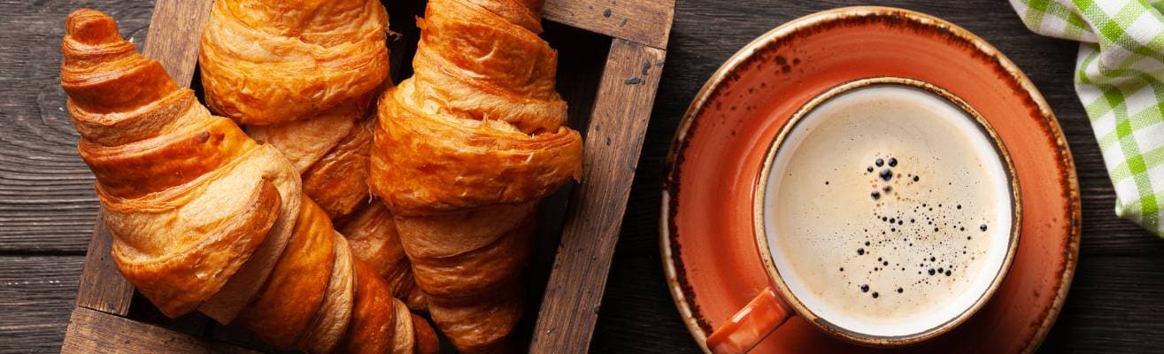 Fresh croissants in a wooden tray next to a ceramic mug of frothy coffee on a rustic wooden table.